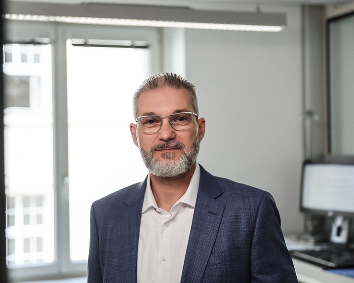 Auf dem Bild sehen wir Robert Hanke. Er trägt kurze grau melierte kurze Haare einen Vollbart und eine Brille. Er steht in einem Büro und lächelt in die Kamera. Robert trägt ein weißes Hemd und einen dunkelblauen Anzug.